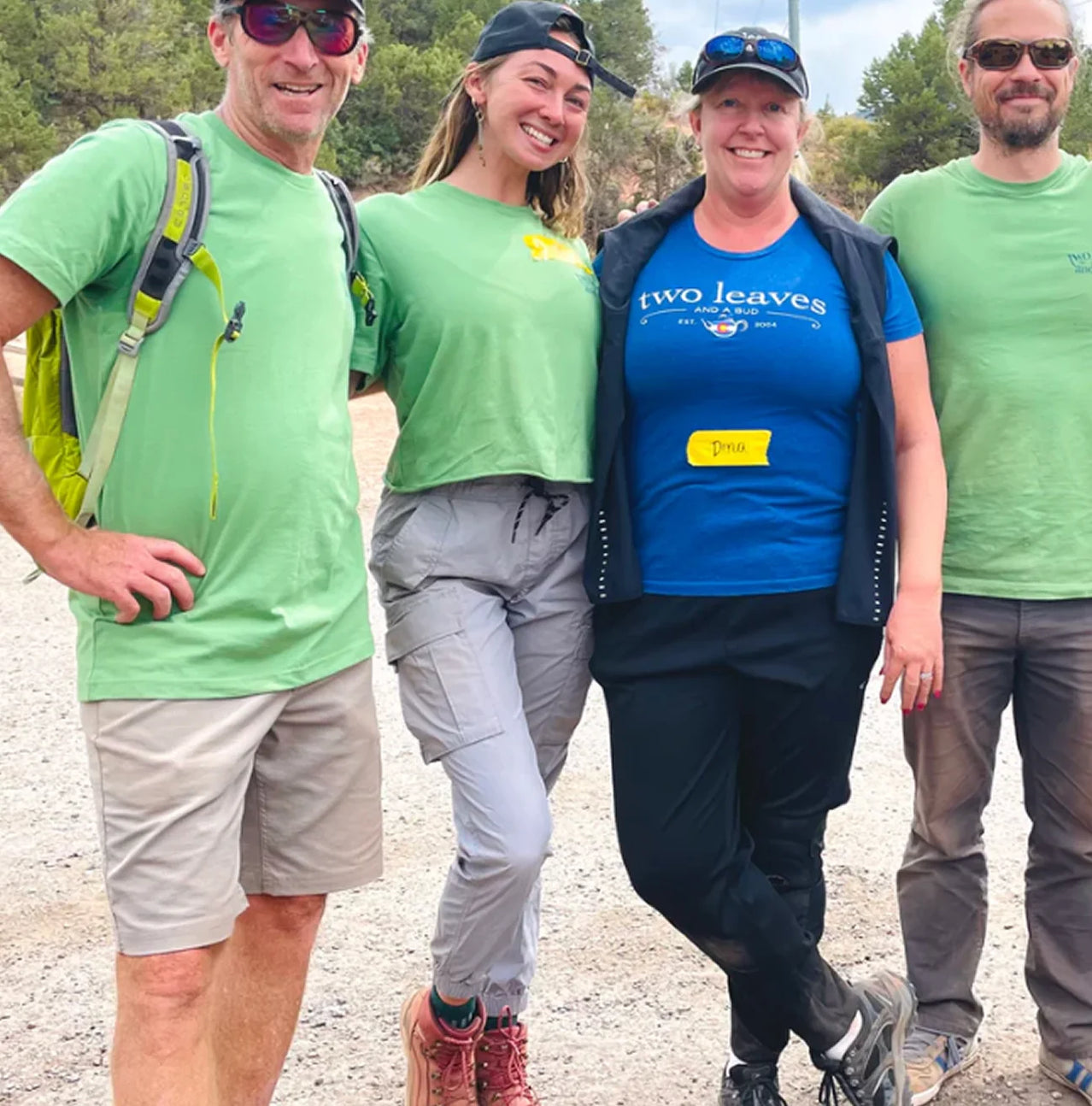Four people posing together outdoors, three in green shirts and one in a blue shirt with 'Two Leaves' logo.