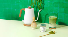 Matcha preparation setup with a kettle, container, and glass on a green tiled wall background.