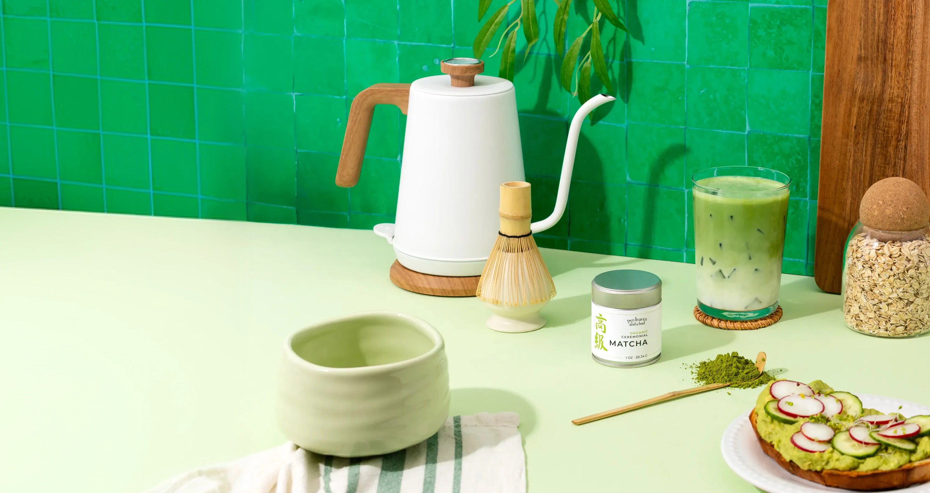Matcha tea setup with a kettle, bowl, and green tea on a light green surface.